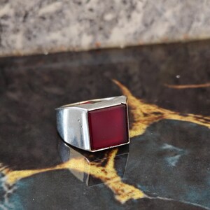 May include: A silver-toned ring featuring a smooth, square-shaped deep red stone set into the center of the band. The metal band has a polished finish and a wide, solid construction that tapers slightly toward the back of the ring.