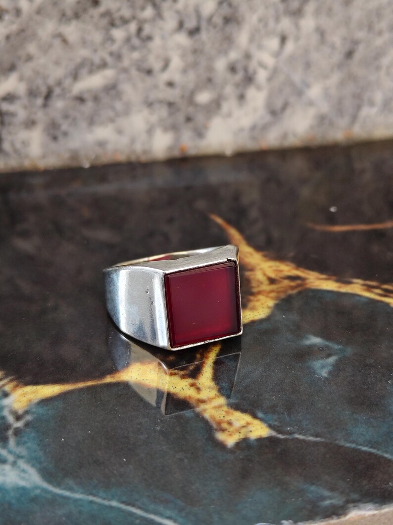 May include: A silver-toned ring featuring a smooth, square-shaped deep red stone set into the center of the band. The metal band has a polished finish and a wide, solid construction that tapers slightly toward the back of the ring.