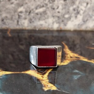May include: A silver-toned metal ring featuring a prominent, flat, square-shaped deep red stone set into the center of the band. The metal band has a smooth, polished finish with a wide, solid construction that tapers slightly toward the back.