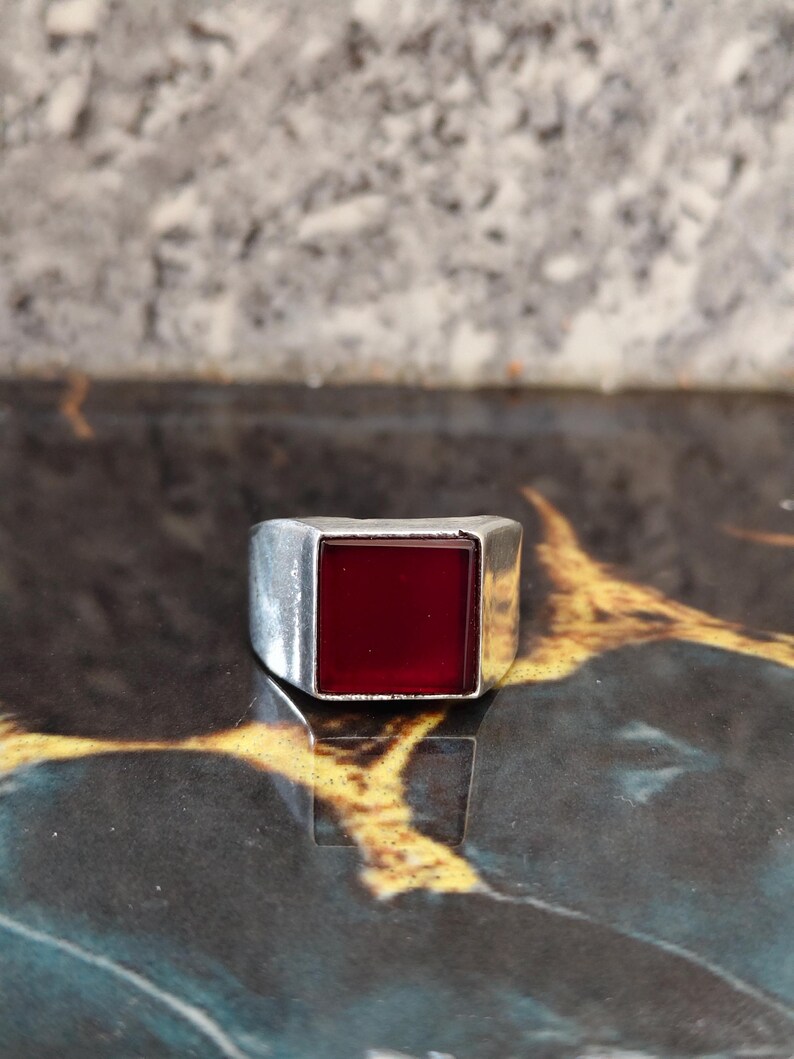 May include: A silver-toned metal ring featuring a prominent, flat, square-shaped deep red stone set into the center of the band. The metal band has a smooth, polished finish with a wide, solid construction that tapers slightly toward the back.