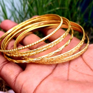 Può includere: Un set di quattro braccialetti rigidi color oro con texture varie. Un bracciale presenta una superficie a perline, mentre gli altri mostrano complessi motivi floreali e geometrici incisi. Questi braccialetti circolari hanno una finitura lucida.