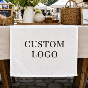 Puede incluir: Un camino de mesa rectangular de tela blanca con el texto negro CUSTOM LOGO centrado en la parte frontal. El camino está colocado sobre un mantel de color beige claro en una mesa de madera, debajo de una jarra blanca y una cesta de mimbre.