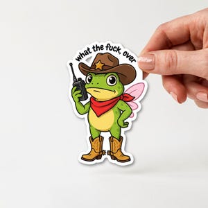 Puede incluir: Una pegatina troquelada que muestra una rana verde con un sombrero de vaquero marrón con una estrella dorada, un pañuelo rojo y botas de vaquero marrones con espuelas. La rana sostiene un walkie-talkie negro y tiene alas rosas. El texto dice what the fuck, over.