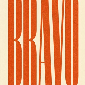 Puede incluir: Un póster vertical que presenta la palabra BRAVO en letras mayúsculas grandes, negritas, de color naranja y sin remates sobre un fondo crema. Debajo del texto principal, la frase A MOMENT WORTH MARKING está impresa en mayúsculas negras más pequeñas.