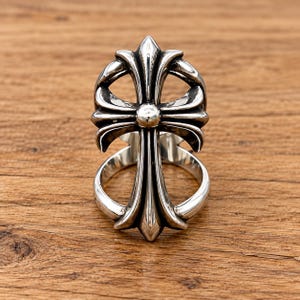 Puede incluir: Un anillo de tono plateado pulido que presenta un diseño de cruz grande y ornamentada con brazos curvos y acampanados. La cruz está centrada en una banda circular, mostrando un acabado metálico con áreas hundidas oscuras que resaltan los contornos.