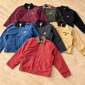 May include: A collection of seven canvas work jackets in various colors including red, blue, black, charcoal, olive green, and tan. Each jacket features a fold-down collar, a full-length front zipper, a zippered chest pocket, and a small square patch.