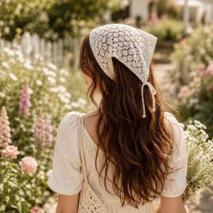 May include: A white triangular headscarf featuring an intricate openwork crochet pattern with a floral motif. The headscarf is tied at the back with two thin crocheted strings that hang down, and it is worn over long brown hair with a white top.