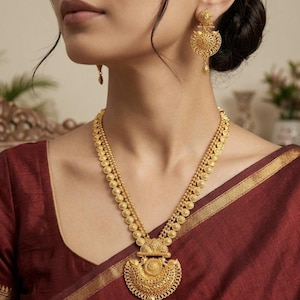 Puede incluir: Un conjunto de collar y pendientes de oro con intrincado trabajo de filigrana. El collar consta de dos cadenas adornadas con pequeños discos dorados, que conducen a un gran colgante semicircular con detalles metálicos y una pequeña gota.