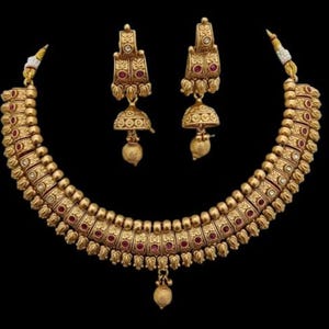 Puede incluir: Un collar y un par de pendientes a juego en tono dorado con intrincados patrones en relieve, pequeñas piedras rojas incrustadas y colgantes en forma de campana. El collar presenta un diseño curvo y segmentado con un dije central.