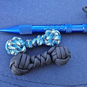 Può includere: Una penna tattica in metallo blu brillante posizionata sopra due nodi a pugno di scimmia in paracord. Un nodo presenta un motivo mimetico blu, nero e bianco, mentre l'altro nodo è nero tinta unita. Entrambi i nodi sono collegati da un breve cordino.