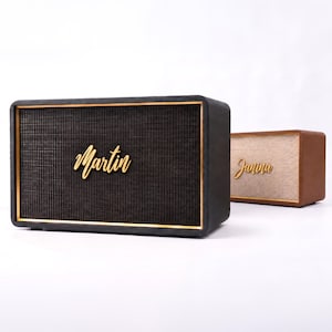 May include: Two rectangular speakers with gold trim and gold script lettering. The front speaker is black with the name Martin, and the rear speaker is brown with the name Janina. Both feature a textured mesh grille and a solid, boxy frame.