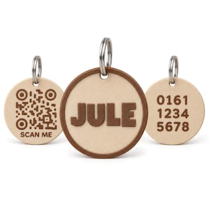 Könnte beinhalten: Drei runde Haustiermarken aus hellem Holz mit Metallringen. Die mittlere Marke zeigt den Namen JULE in Braun. Die linke Marke enthält einen braunen QR-Code mit dem Text SCAN ME. Die rechte Marke zeigt die braune Zahlenfolge 0161 1234 5678.