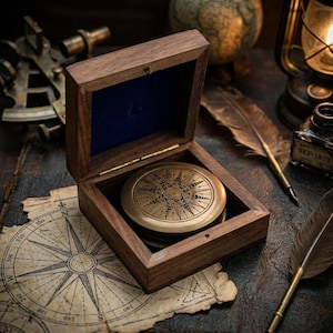 Peut inclure: Une boussole ronde en laiton avec un motif détaillé en forme d'étoile est placée dans une boîte carrée en bois ouverte. La boîte présente une finition en bois sombre et une doublure intérieure en velours bleu profond sur du papier parchemin.
