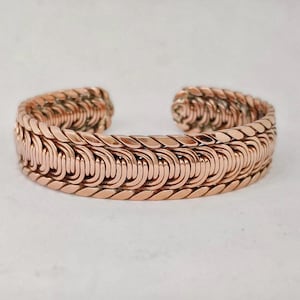 May include: A copper-colored cuff bracelet featuring a complex, woven wire design. The bracelet is constructed from multiple strands of metal wire twisted and looped together to create a repeating, textured pattern with an open back design.