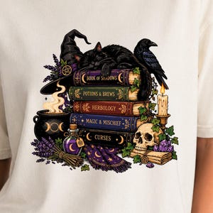 May include: An ivory t-shirt featuring a graphic of a black cat and raven resting on a stack of books titled Book of Shadows, Potions & Brews, Herbology, Magic & Mischief, and Curses, alongside a cauldron, skull, candle, and lavender sprigs.