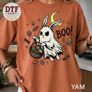 May include: A rust-colored t-shirt featuring a graphic of a white ghost rabbit holding a basket filled with colorful eggs and candy. The design includes a yellow crescent moon, scattered stars, and the text BOO! in a black, jagged font.