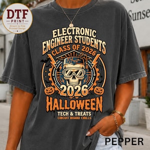 May include: A charcoal gray t-shirt featuring a graphic print with a skull wearing a graduation cap and goggles, surrounded by gears, lightning bolts, and pumpkins. The text reads Electronic Engineer Students Class of 2026 Halloween Tech & Treats.