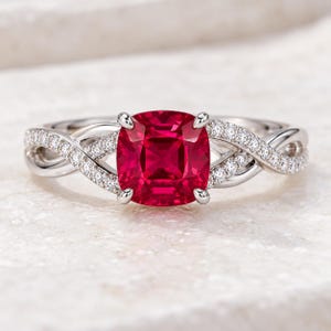 Puede incluir: Un anillo de tono plateado con una piedra preciosa roja intensa de corte cojín sujeta por cuatro garras. La banda presenta un intrincado diseño trenzado, con las secciones exteriores incrustadas de pequeñas piedras claras y brillantes.