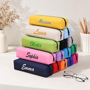 Peut inclure: Une pile de trousses rectangulaires de différentes couleurs unies, notamment jaune, crème, vert, rose et bleu marine. Chaque trousse possède une fermeture éclair noire et est brodée d'un prénom : Mason, Luna, Olivia, Sophia ou Emma.