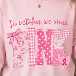 Könnte beinhalten: Ein hellrosa Langarm-Sweatshirt mit dem Text In october we wear PINK in Schreibschrift. Die Buchstaben sind mit verschiedenen rosa Mustern gefüllt, darunter Vichy-Karo, Punkte und Blumenmuster, sowie einem rosa Herz-Symbol mit Schleife.