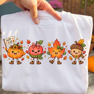 May include: A white t-shirt featuring a colourful graphic of four anthropomorphic autumn characters: a pumpkin holding a sign that says Hello Fall, an apple, a maple leaf, and an acorn. Each character wears boots, gloves, and seasonal accessories.