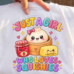 May include: A white t-shirt featuring a colorful graphic with the text JUST A GIRL WHO LOVES SQUISHIES. The design includes a cute dumpling in a bamboo steamer, a jar of strawberry jam, and a block of butter, all with smiling faces.