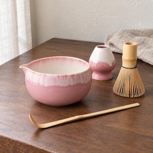 May include: A ceramic matcha bowl with a pink and white gradient glaze, a matching pink and white ceramic whisk holder, a bamboo whisk, and a long bamboo scoop. The bowl features a small pouring spout on one side and a smooth, rounded shape.