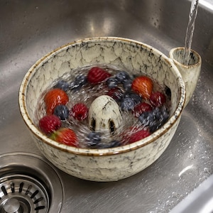 May include: A cream-colored ceramic berry colander with a dark crackle glaze pattern, featuring a central drainage cone and a side handle. Fresh red strawberries and dark blue blueberries are being rinsed with water inside the bowl.