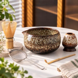 May include: A matcha tea set including a bamboo whisk, a ceramic bowl with a brown and cream mottled glaze, a matching whisk holder, a stainless steel mesh strainer, and a bamboo scoop, all arranged on a white surface.