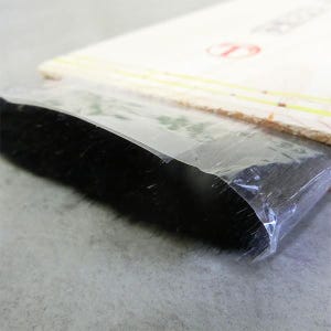 May include: A flat brush with dense black bristles encased in a clear plastic protective sleeve. The handle is made of light-coloured wood and features a red circular logo with a letter T inside, visible near the top edge of the wooden handle.
