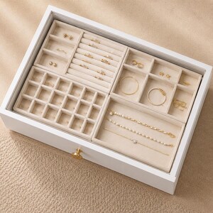 May include: A white wooden jewellery organiser drawer with a glass top and a gold knob. The interior features beige velvet inserts divided into sections for rings, earrings, bracelets, and necklaces, containing various gold-toned jewellery pieces.