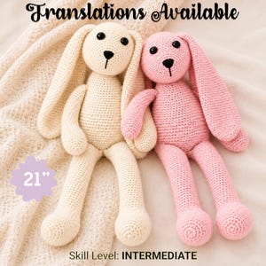 Op de afbeelding: Twee gehaakte konijnenpoppen, een crème en een roze, elk met lange oren, zwarte knoopogen en een geborduurde neus. Een paarse afbeelding links toont de tekst 21 inches. De tekst Translations Available en Skill Level: INTERMEDIATE is zichtbaar.