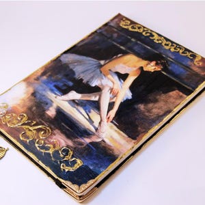 May include: A rectangular hardcover journal featuring a printed painting of a ballerina in a white tutu on the front cover. The cover is accented with raised gold filigree detailing along the edges and includes a gold-toned metal ballerina charm.