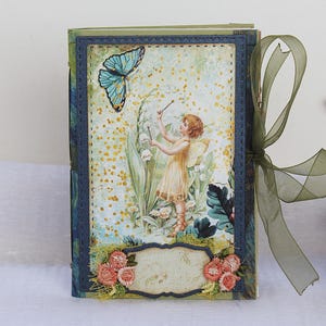 Puede incluir: Un diario rectangular con un marco de borde azul oscuro cosido. La portada presenta una ilustración central de un hada con vestido amarillo entre flores y una mariposa azul. La parte inferior incluye una etiqueta en blanco con detalles florales rosas.