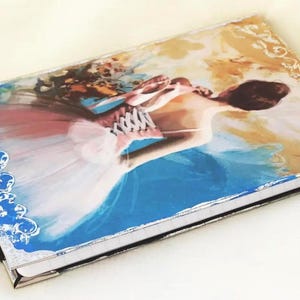 May include: A rectangular hardcover journal featuring a printed illustration of a person in a pink tutu and corset. The cover has blue and gold decorative borders, silver-toned metal corner protectors, and a black ribbon bookmark with a metal charm.