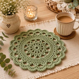 May include: A round, sage green crocheted doily featuring an intricate, radiating floral pattern with a scalloped edge. The textile is crafted with a dense, textured yarn, showcasing a symmetrical design that expands from the center outward.