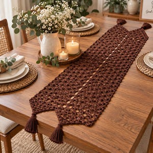 Può includere: Un lungo runner da tavola rettangolare all'uncinetto color marrone scuro, caratterizzato da un intricato motivo traforato e rifinito con quattro nappe abbinate agli angoli. Il runner è disposto nel senso della lunghezza su un tavolo da pranzo.