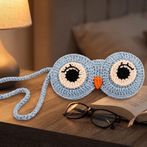 Op de afbeelding: Een lichtblauw gehaakt slaapmasker in de vorm van een uil, voorzien van twee grote ronde ogen met witte en zwarte garendetails, een kleine oranje snavel en een bijpassend lichtblauw gehaakt bandje om het masker rond het hoofd te bevestigen.