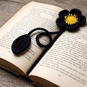 May include: A handmade crochet bookmark featuring a black five-petaled flower with a bright yellow center, attached to a long black cord that ends in a single black leaf-shaped accent, resting between the pages of an open hardcover book.