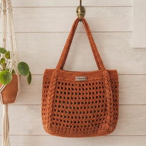 Puede incluir: Un bolso tote de ganchillo en color naranja quemado con un patrón de rejilla calado, dos asas trenzadas y una pequeña etiqueta metálica rectangular centrada cerca de la abertura superior. El bolso cuelga de un gancho en una pared de madera clara.