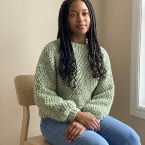 May include: A sage green long-sleeve sweater with a textured, chunky knit pattern, featuring a ribbed crew neckline and ribbed cuffs. The sweater is paired with light blue denim jeans, creating a casual and simple outfit combination.