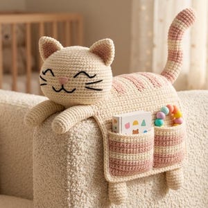 May include: A cream-colored crochet cat organizer with pink striped accents on the back and tail. The cat features embroidered black eyes and whiskers, a pink nose, and two front pockets containing a small white book and a colorful bead toy.