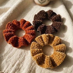 May include: Three circular hair scrunchies made of a textured, plush velvet fabric. The set includes one in a deep burnt orange, one in a dark chocolate brown, and one in a warm golden mustard yellow color, each featuring a gathered, ruffled design.