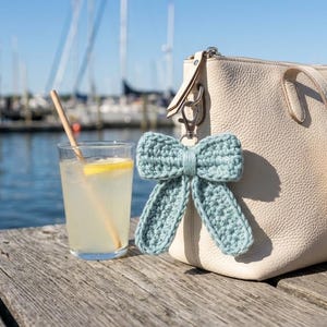 Puede incluir: Un llavero de lazo tejido a ganchillo en color azul claro sujeto a un bolso de cuero color crema, colocado junto a un vaso transparente con una bebida amarilla, una rodaja de limón y una pajita de madera, frente a un puerto con barcos.