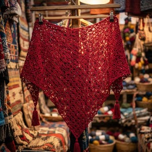 Può includere: Uno scialle triangolare rosso vivace caratterizzato da un intricato motivo a uncinetto traforato. Il capo è appeso a una gruccia di legno e presenta tre nappe rosse abbinate fissate a ciascuno dei tre angoli del design triangolare.
