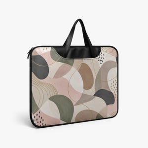 Puede incluir: Una funda rectangular para portátil con asas y bordes negros, que presenta un patrón abstracto de formas orgánicas superpuestas en tonos apagados de verde salvia, rosa empolvado, beige y gris carbón, acentuado con finas líneas doradas y puntos negros.