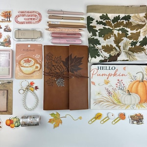 May include: A collection of autumn-themed stationery including a brown leather-style journal with leaf prints, a pouch with a pumpkin graphic and the text Hello Pumpkin, pens, highlighters, stickers, a pearl keychain, and leaf-shaped paper clips.