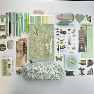 May include: A collection of green-themed stationery items including a floral patterned notebook, a matching pencil case, various pens, bookmarks, stickers, a reading list, a small candle, and decorative paper clips arranged on a flat surface.