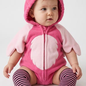 Peut inclure: Une barboteuse à capuche rose vif sur le thème du flamant rose avec une fermeture éclair frontale, des manches en forme d'ailes rose clair et une capuche ornée d'un visage de flamant rose. L'ensemble est porté avec des chaussettes hautes rayées.