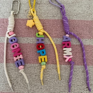 Op de afbeelding: Drie sleutelhangers gemaakt van gekleurd koord en plastic letterkralen. De eerste is wit met het woord JOONIE, de tweede is geel met een sterbedel en het woord JHOPE, en de derde is paars met het woord ARMY. Elke hanger heeft een metalen sluiting.